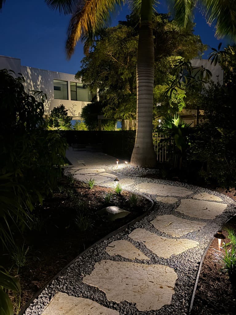 Low-voltage brass walkway lighting leading to a modern luxury coastal residence with illuminated royal palm tree
